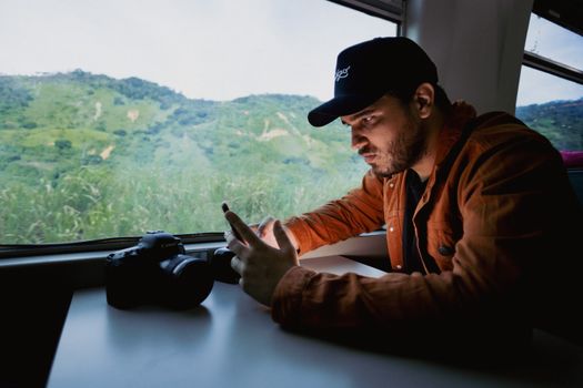 Young man in cap using smartphone by window with camera. Capturing candid travel moments.