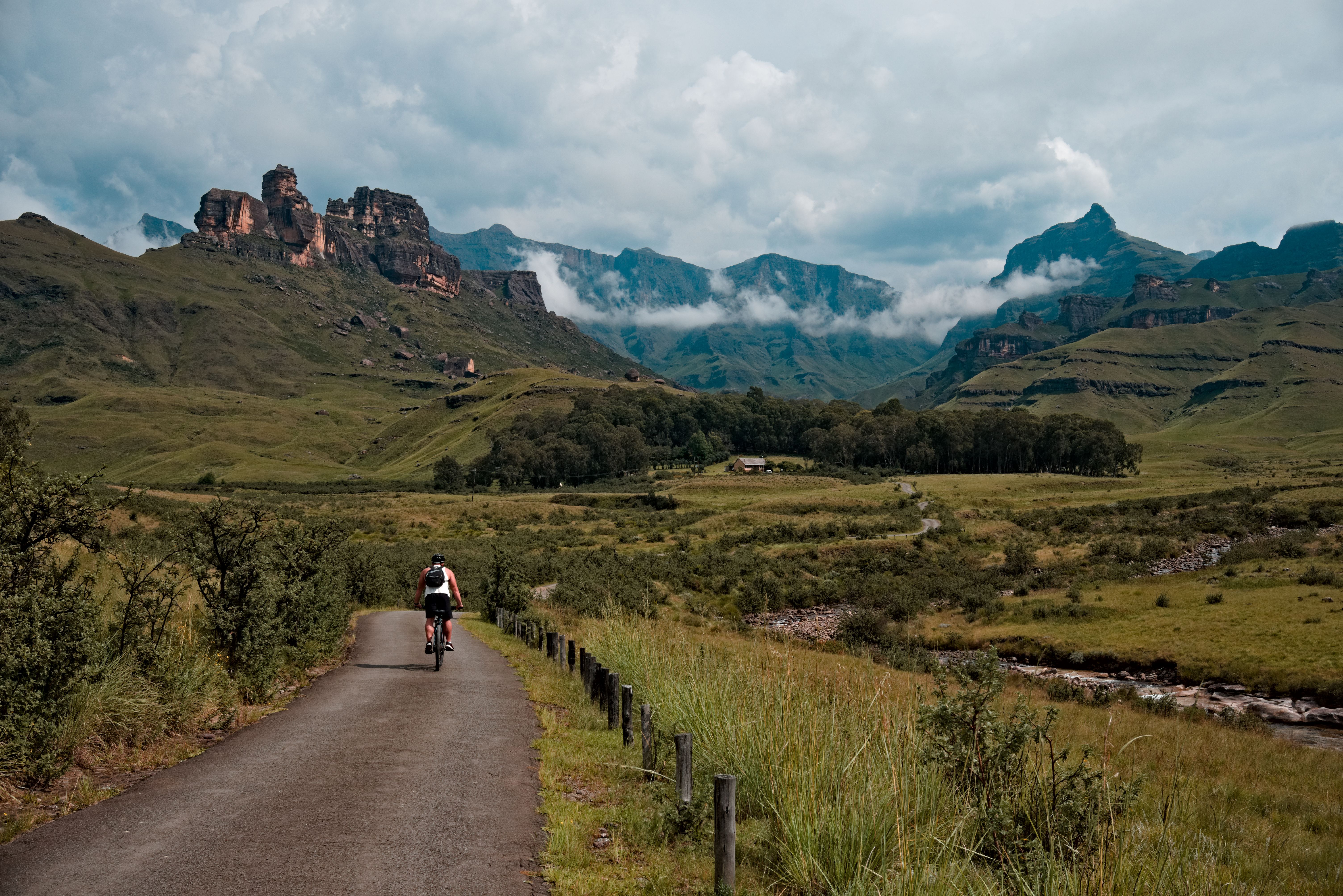 A lone cyclist travels a winding road through lush green valleys and towering mountains.