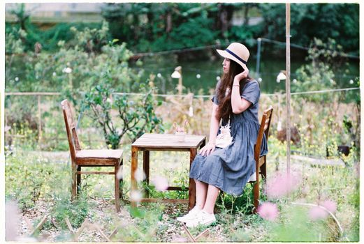 Woman in a Dress and a Hat Sitting by a Table in the Garden