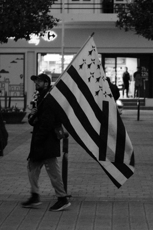 Free A man walks through an urban area carrying the Brittany flag, captured in black and white. Stock Photo