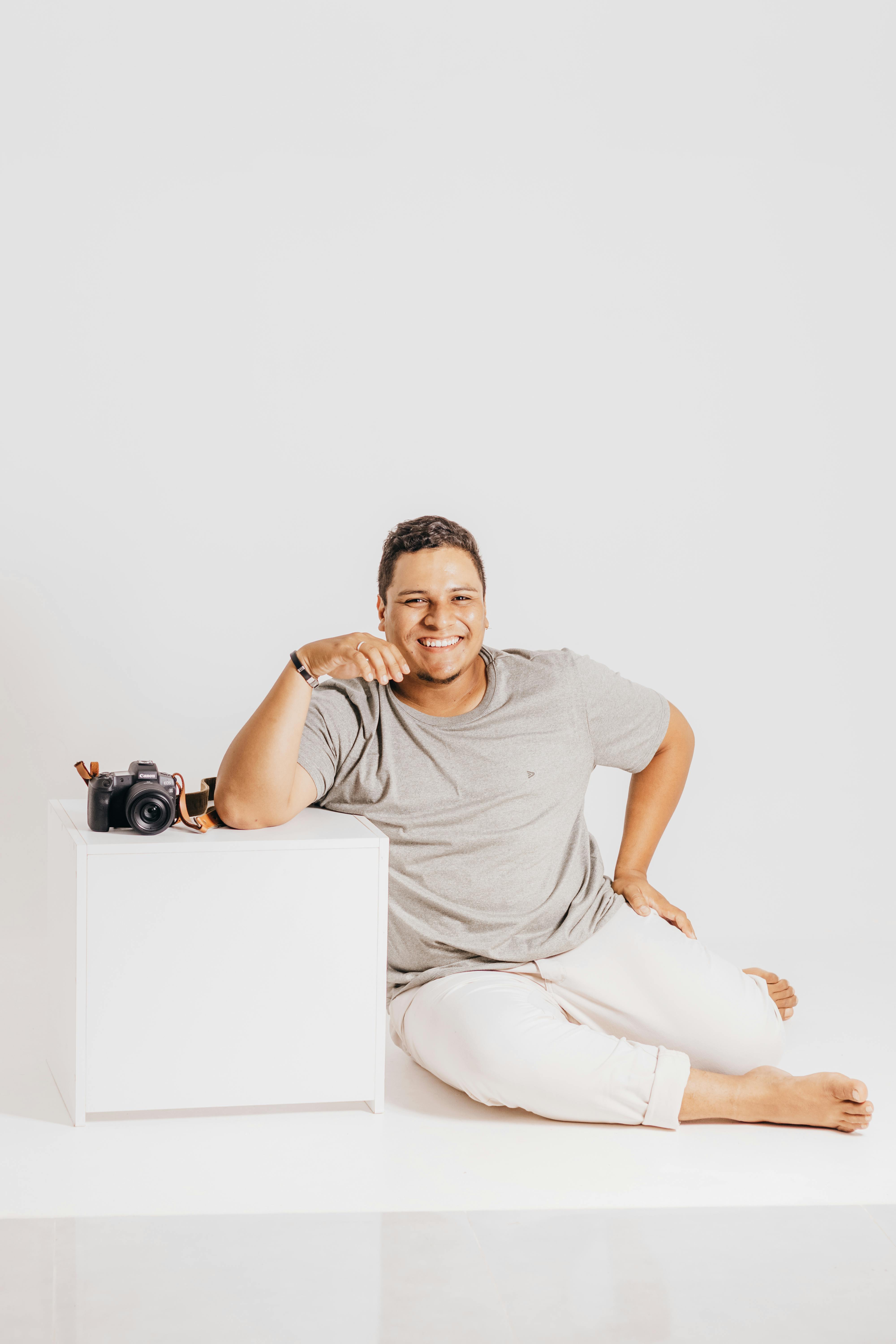 Young man in casual wear sitting on studio floor with a camera on block and white background.