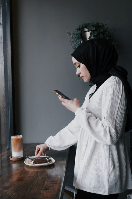 A woman taking a photo of a chocolate cake and coffee at a table using her smartphone.