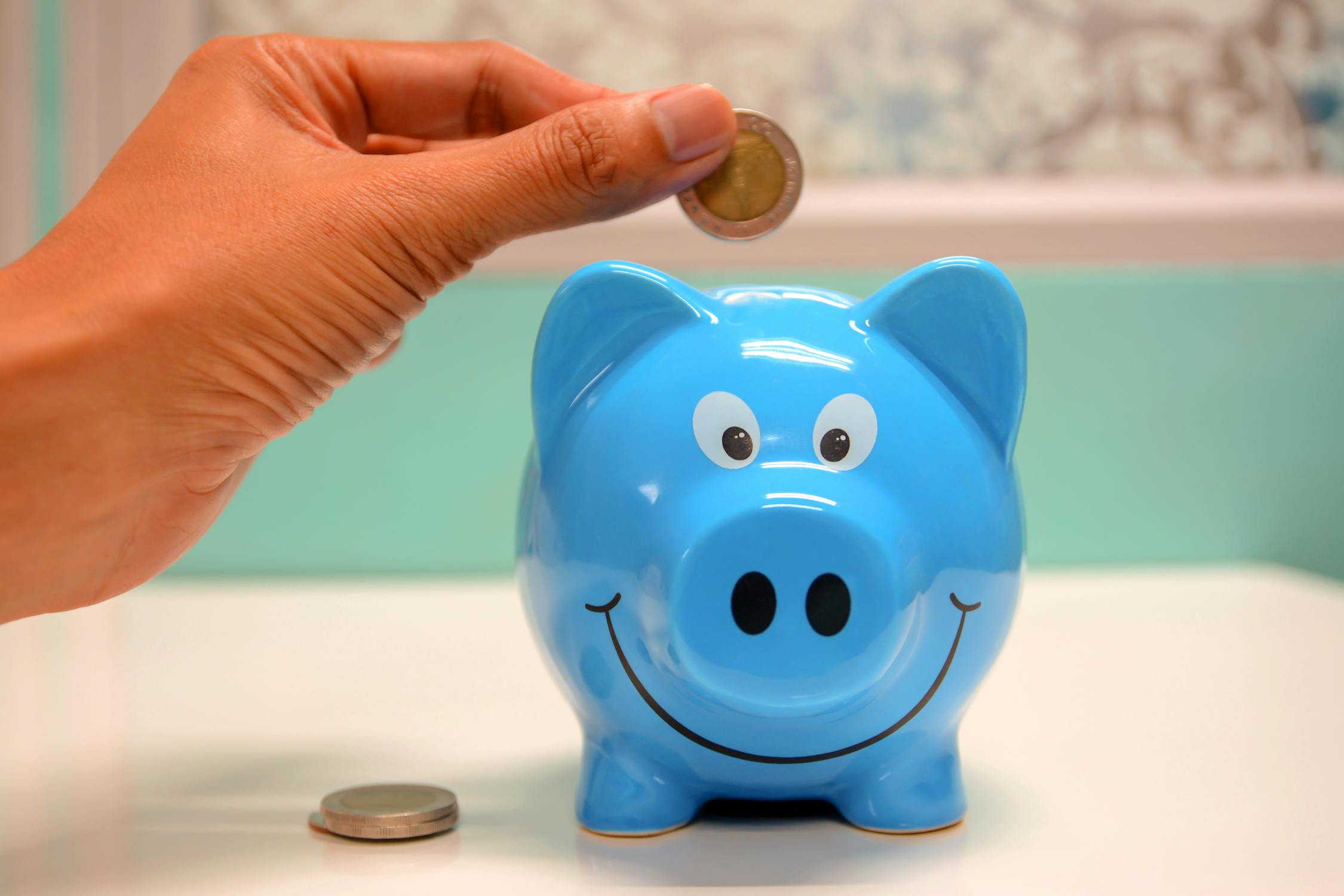 Hand dropping a coin into a blue pig shaped piggie bank with a big smile on its face.