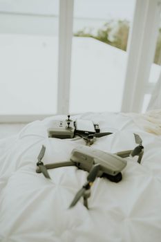 A drone and controller placed on a white bed by a window with a view.