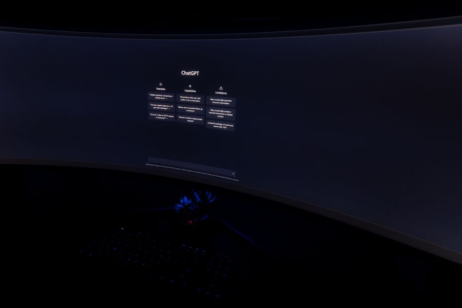 Close-up of a dark room with a curved monitor showing the ChatGPT interface on screen.