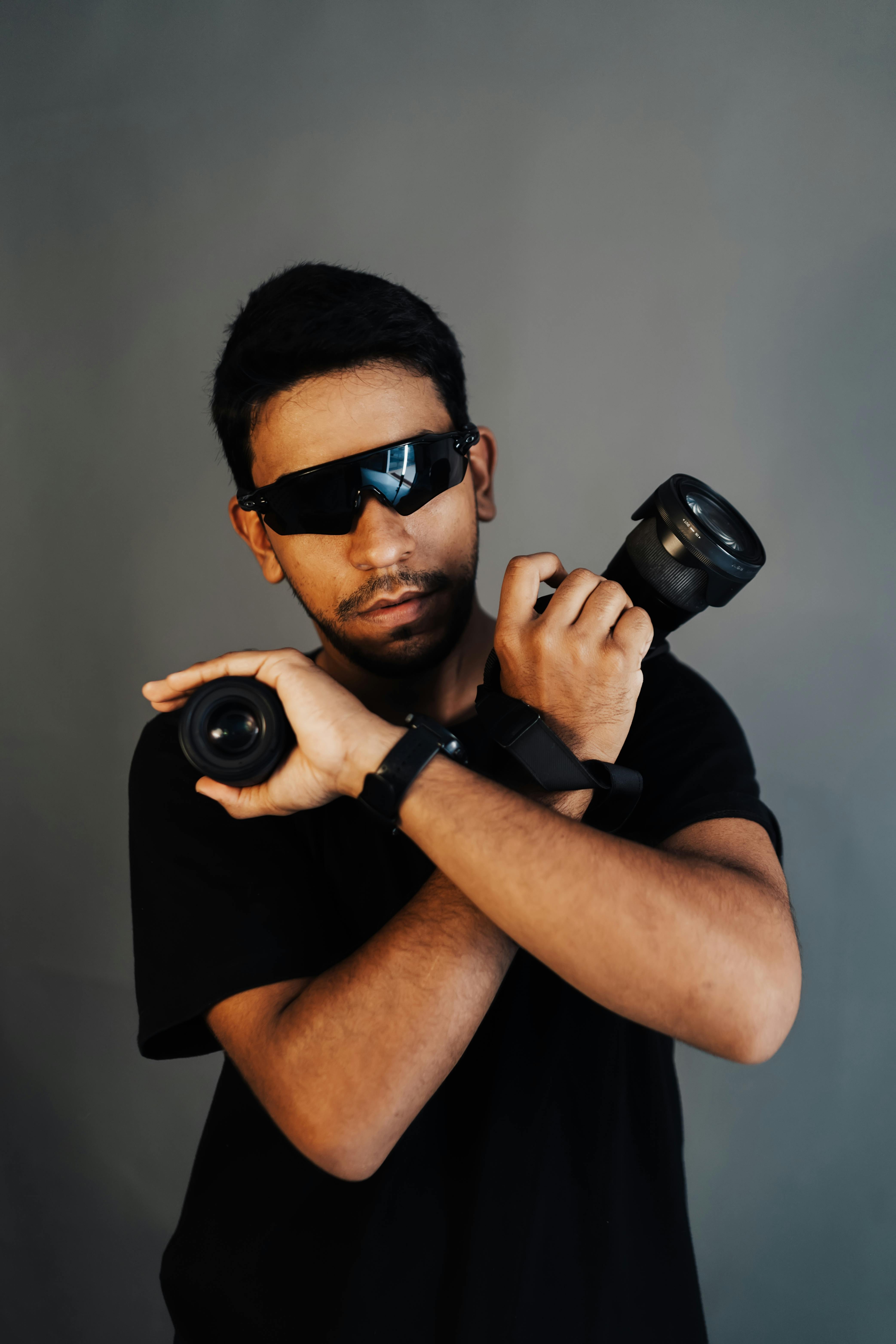 Portrait of a man with sunglasses posing with camera lenses against a gray background.