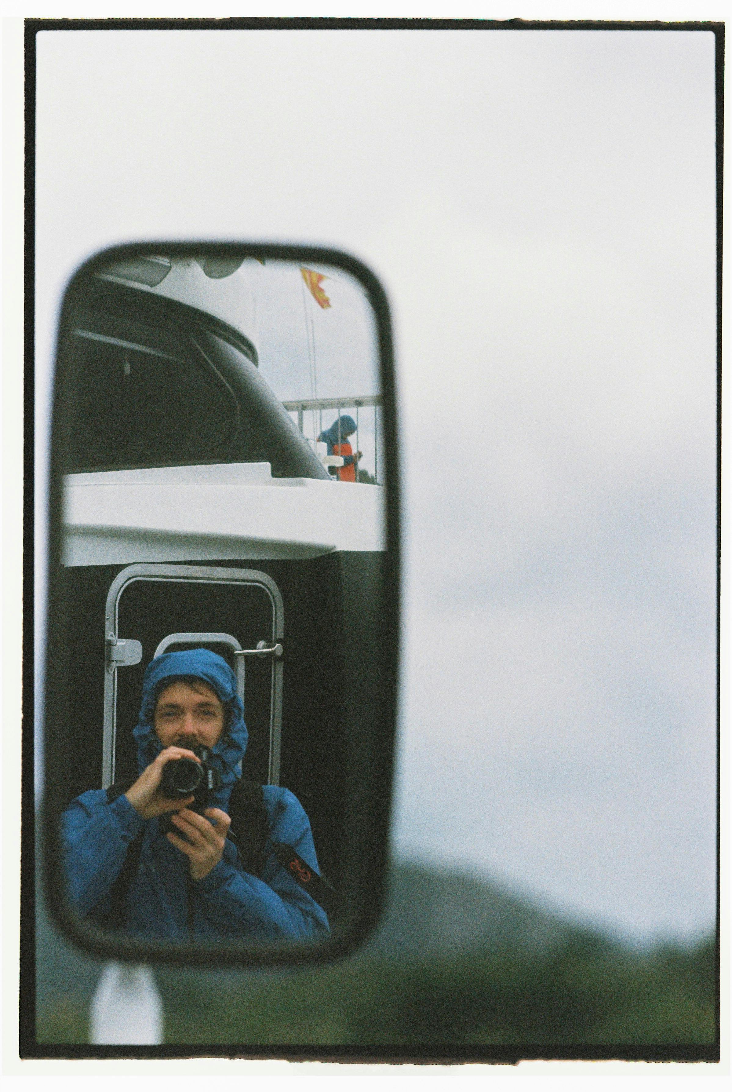 Man in blue jacket capturing photo reflection in vehicle mirror.