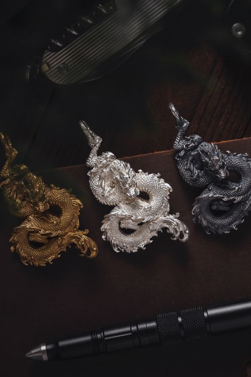Free Three intricately designed dragon pendants in gold, silver, and black alongside a tactical pen on a dark surface. Stock Photo