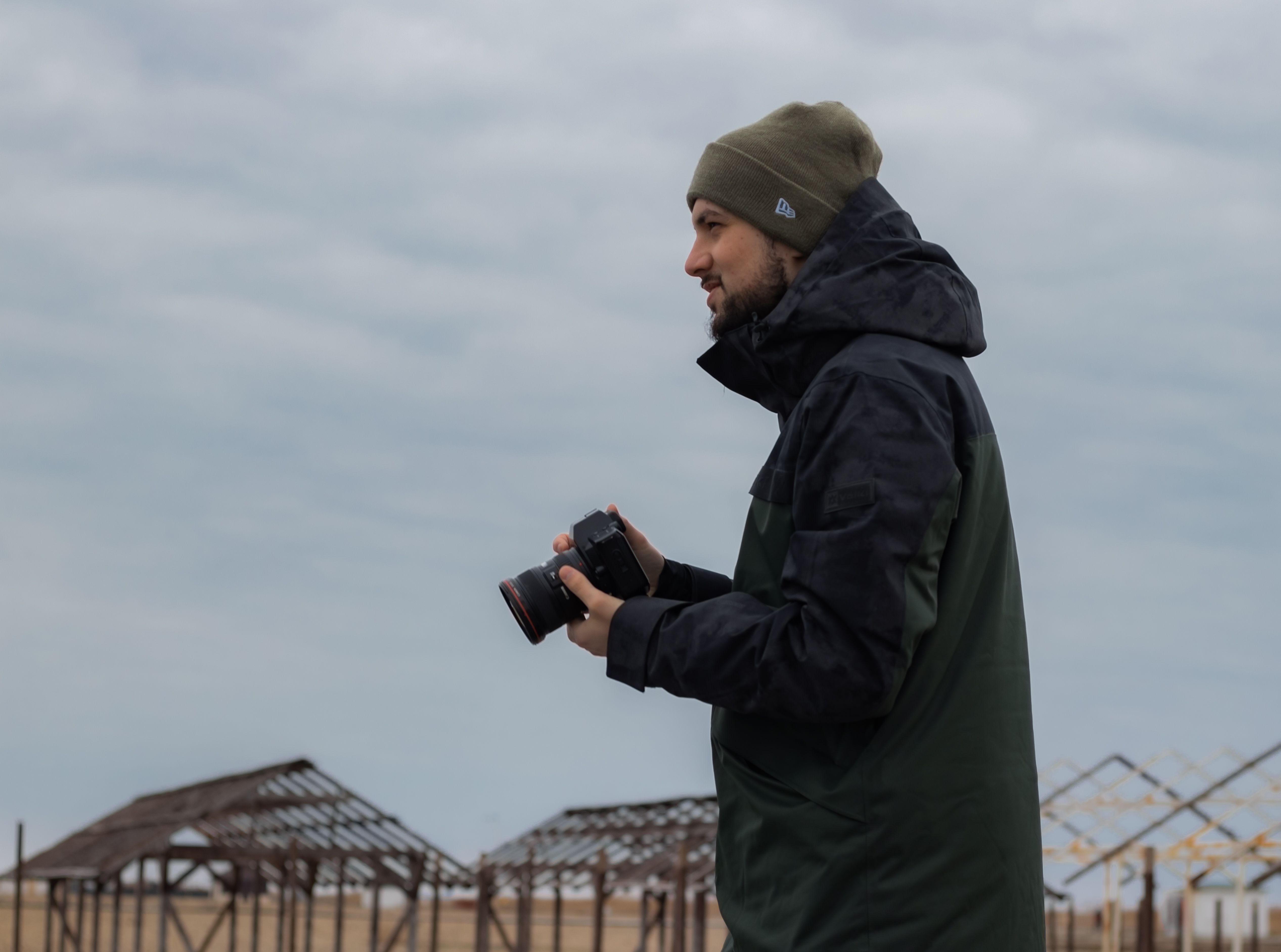 Side profile of a photographer outdoors wearing a beanie and jacket, holding a camera on a cloudy day.