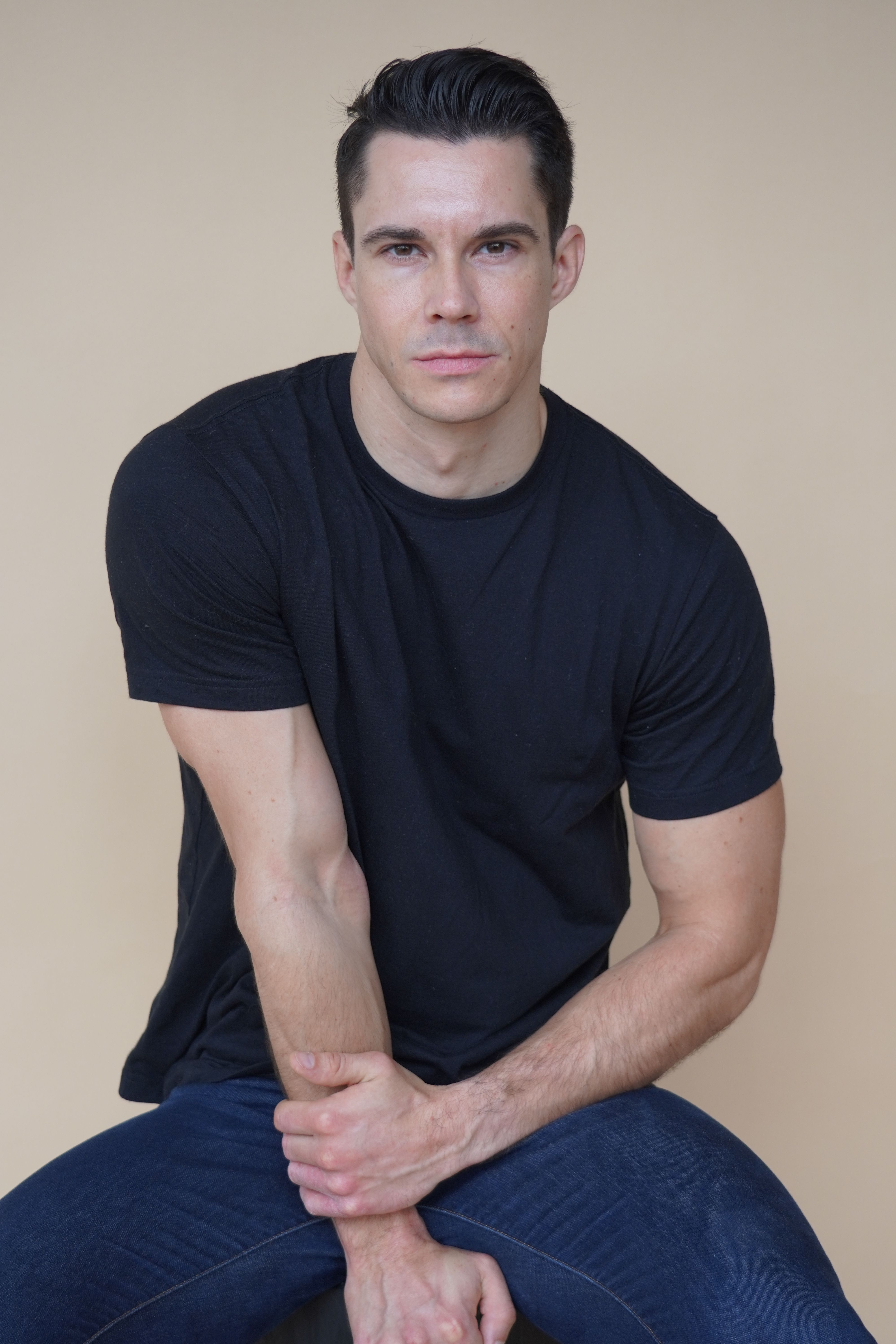 Confident man in jeans and black t-shirt posing elegantly against a neutral background.