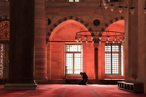 Free A man kneels in prayer inside a beautifully adorned mosque, bathed in warm light from large windows. Stock Photo