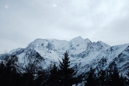 Free Majestic snowcapped mountain range under a cloudy sky, surrounded by winter trees. Stock Photo