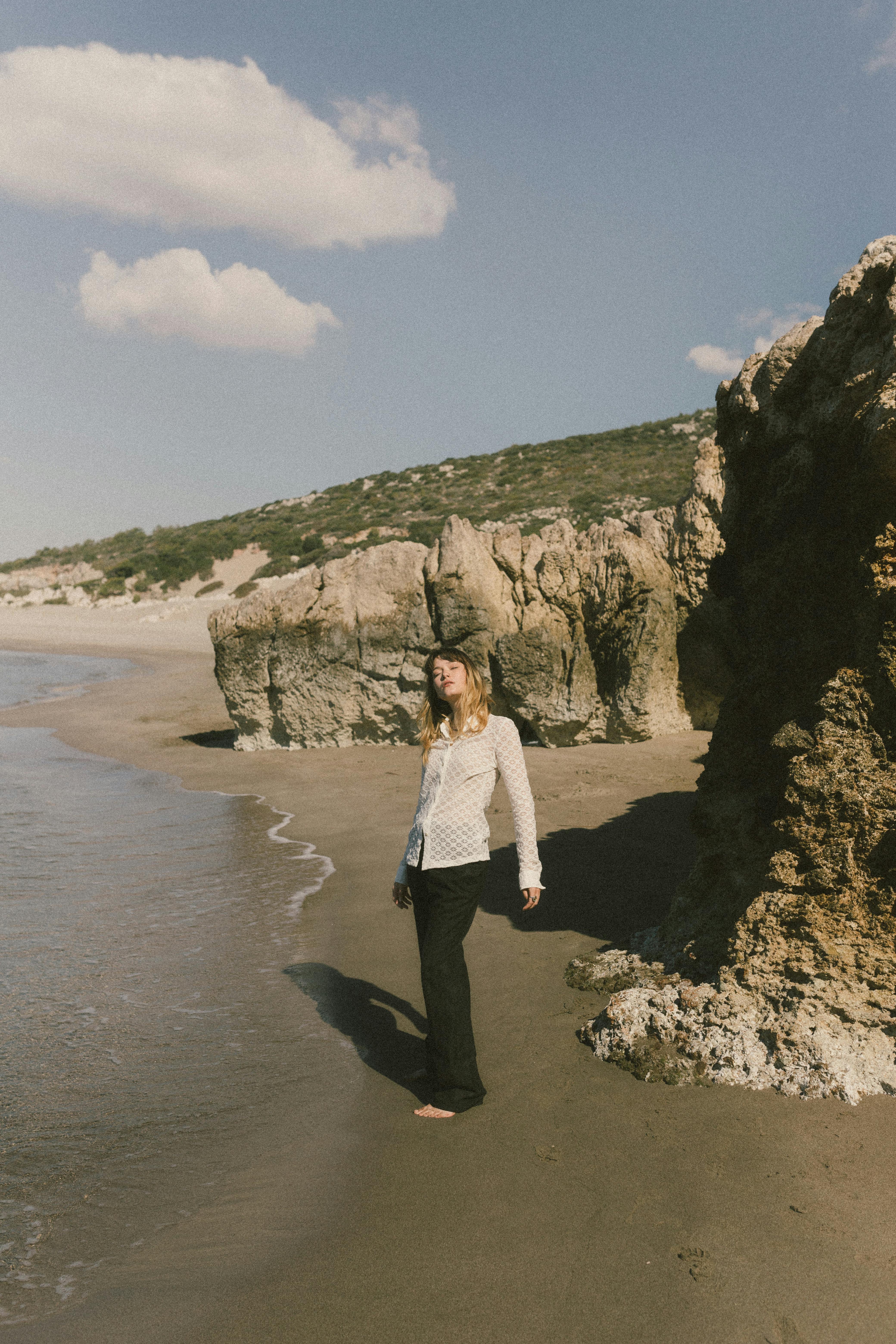 A woman stands barefoot on a sandy beach with rocky cliffs under a clear blue sky.