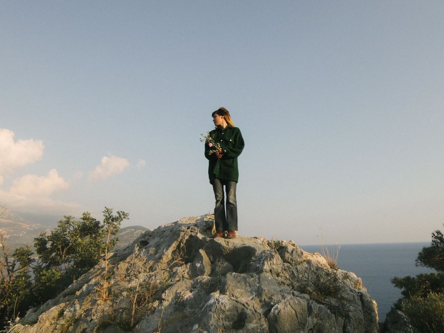 A woman holds flowers while standing on a cliff, gazing at the ocean.