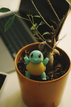 A small blue figurine nestled in a potted plant beside a laptop on a desk.
