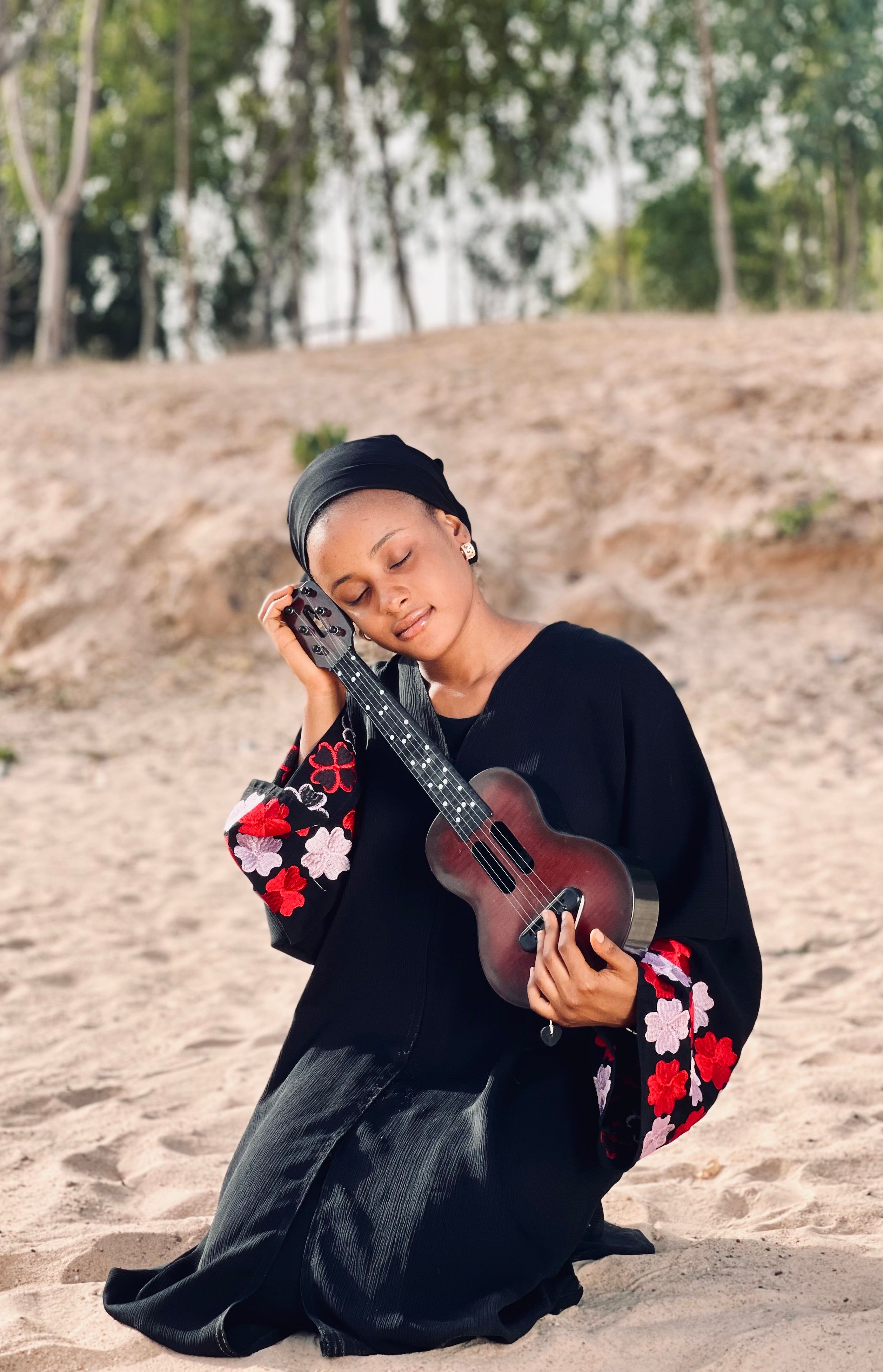 A woman in a floral dress kneels on the beach with a guitar, eyes closed in tranquility.