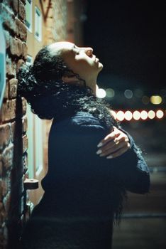 A woman stands in profile against a brick wall in a dimly lit urban setting at night.
