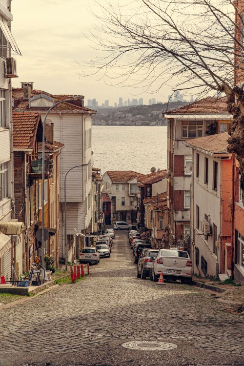 Free Narrow cobblestone street with parked cars leading to a scenic waterfront view. Stock Photo