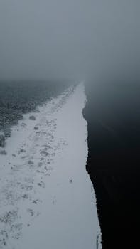 A serene aerial winter landscape showing a snowy forest and river under misty weather in Калнгале, Latvia.