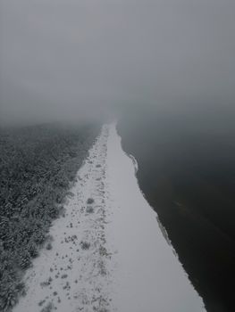 Aerial view of Garciems coastline blanketed in snow and fog, capturing winter's serene beauty.