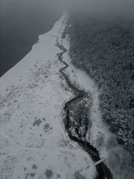A breathtaking aerial shot of a snow-covered winding river beside a lush forest in Mežciems, Latvia.