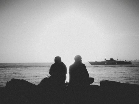 Peaceful silhouette of two people sitting by the sea at sunrise with a ship in the distance.