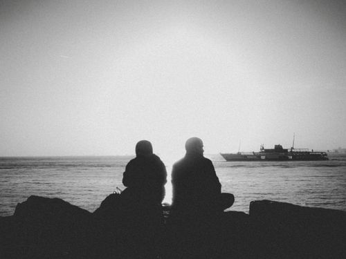 Free Peaceful silhouette of two people sitting by the sea at sunrise with a ship in the distance. Stock Photo