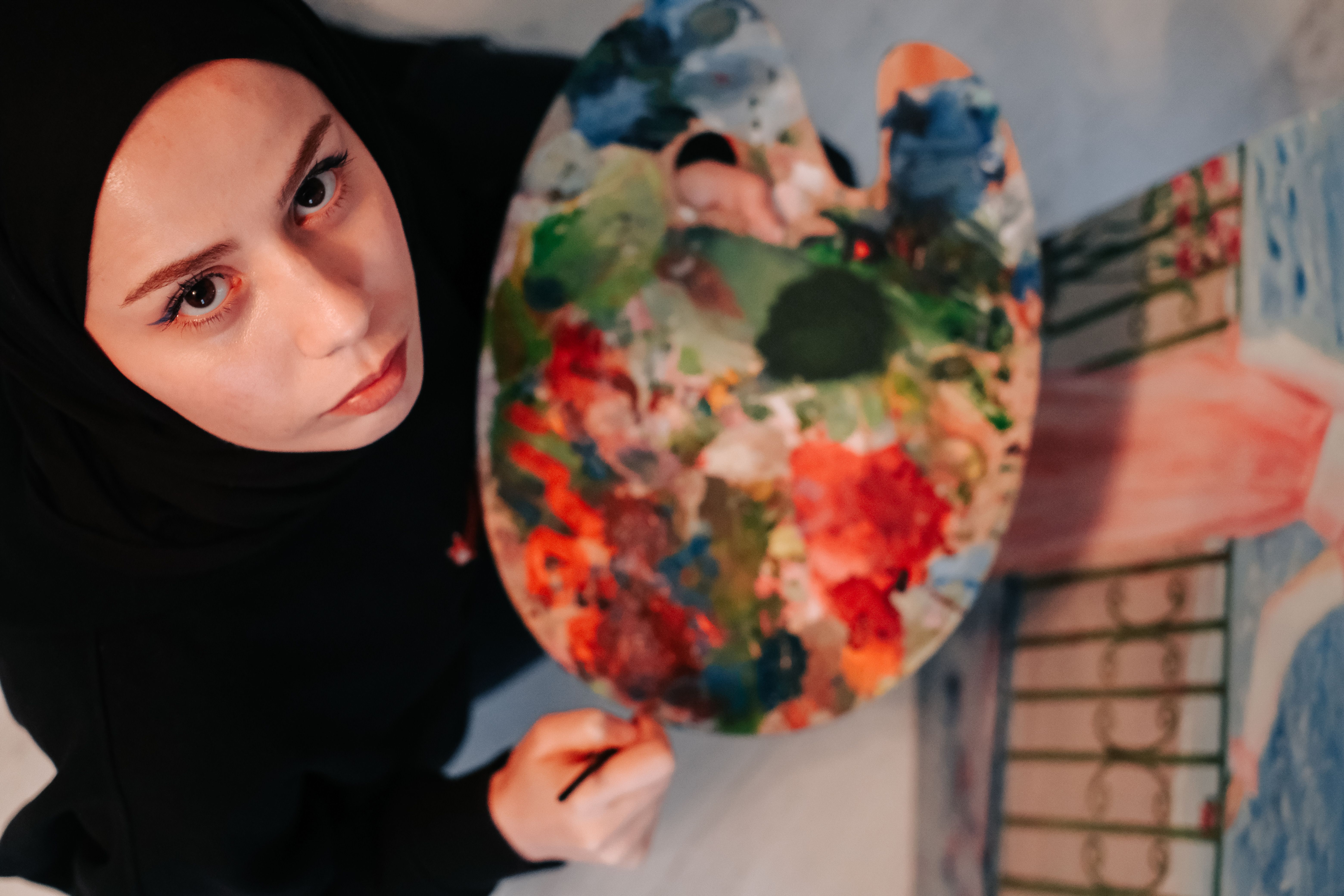 Muslim woman artist with hijab holding colorful paint palette indoors. High angle view.