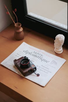 A vintage camera rests on a newspaper by a windowsill, creating a cozy indoor scene with artistic decor.