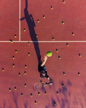 Aerial shot of a tennis player serving on a court surrounded by tennis balls under sunlight.
