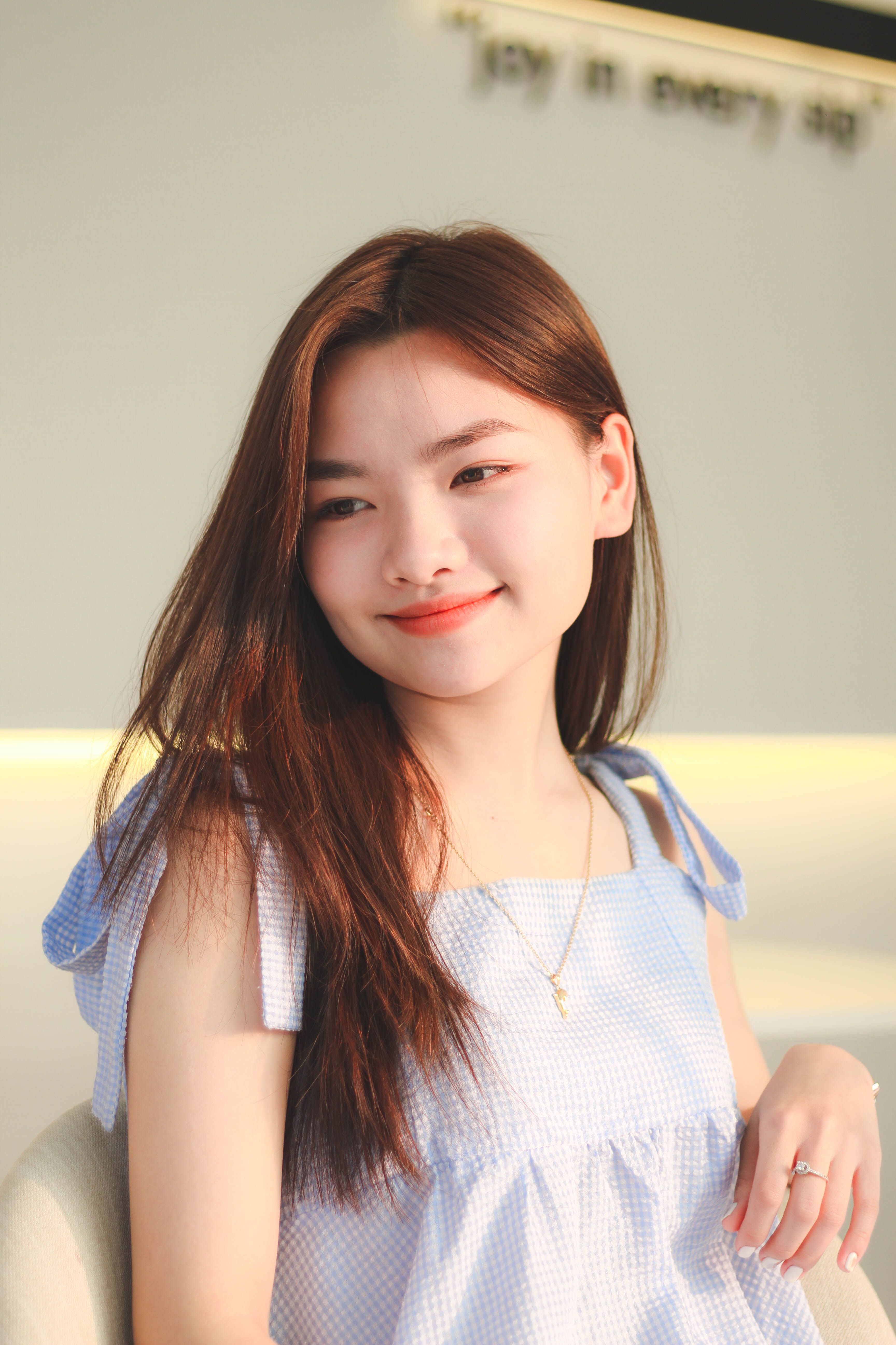 A young woman with long brunette hair smiling indoors in a sunlit environment.