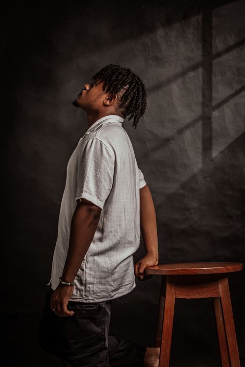 Free Fashion portrait of a young man with dreadlocks and white shirt leaning on a wooden stool in a studio. Stock Photo