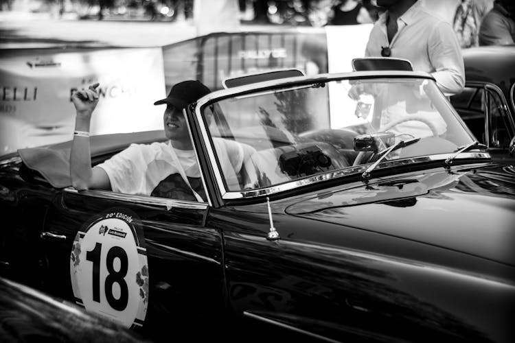 Black And White Photo Of A Man Sitting In A Vintage Car At A Car Show 