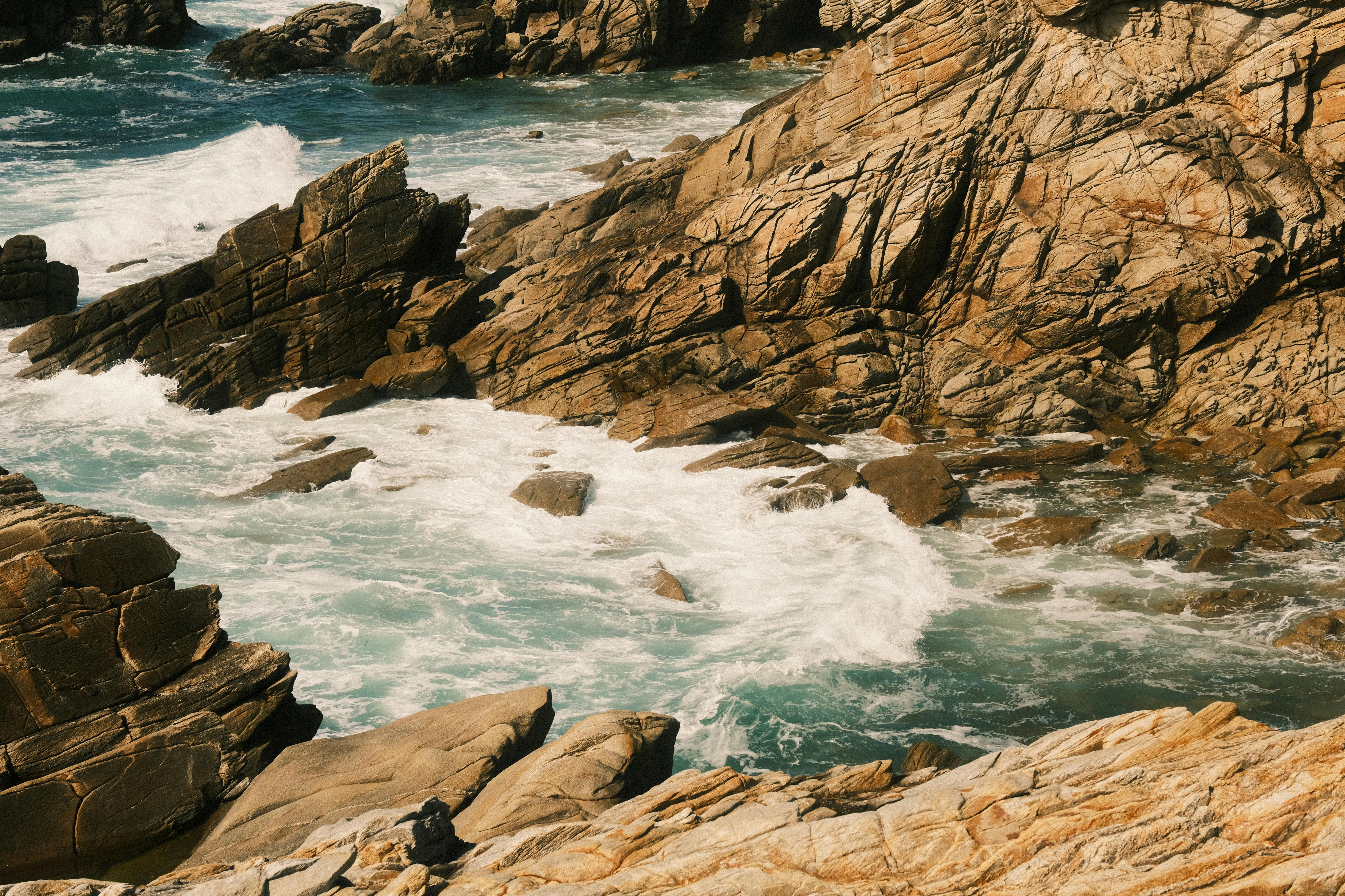 A sunlit rocky coastline showing waves crashing against rugged rocks, creating a dramatic seascape.