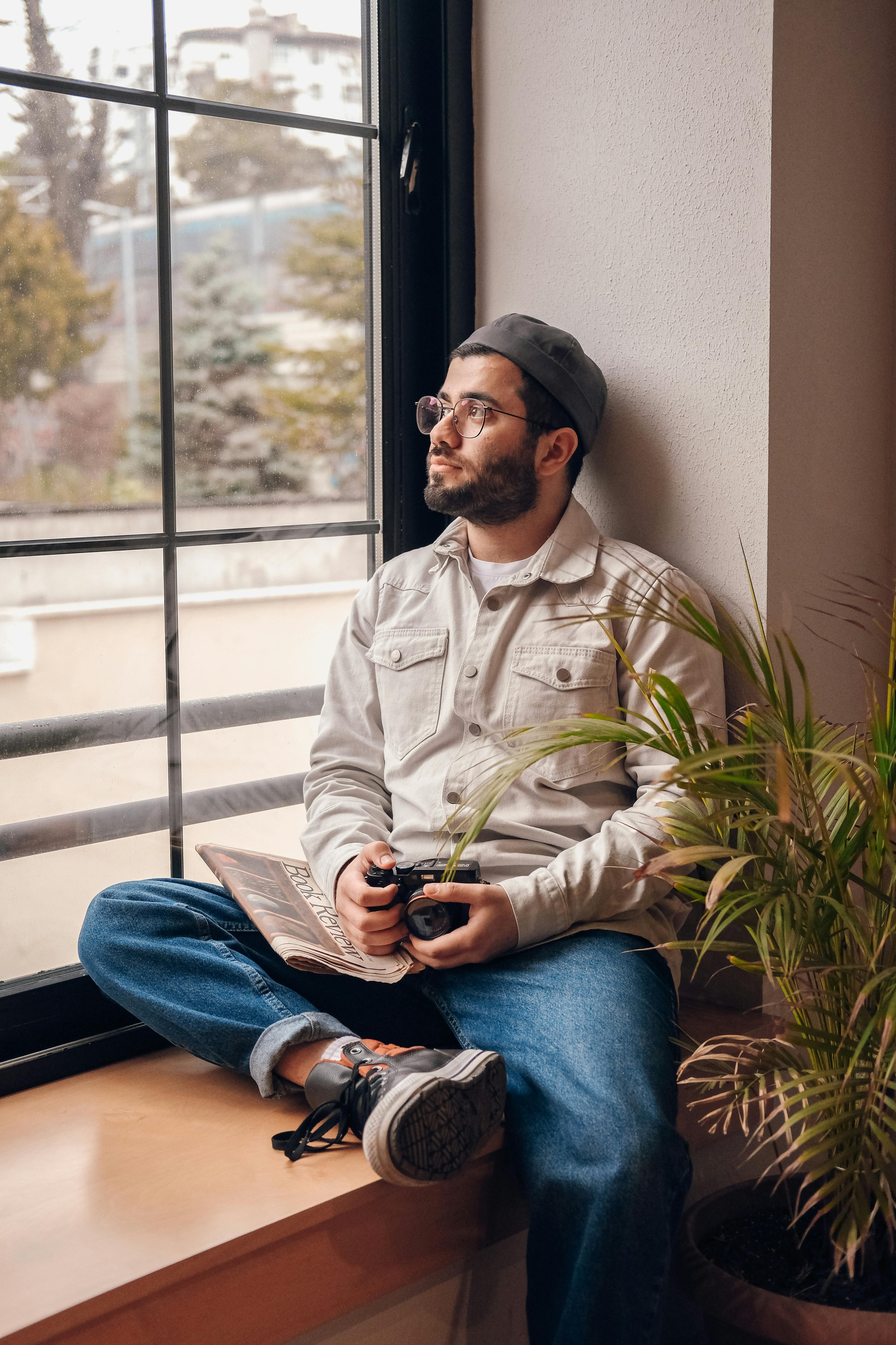 A contemplative man sitting by a windowsill with a camera and book, looking outside.