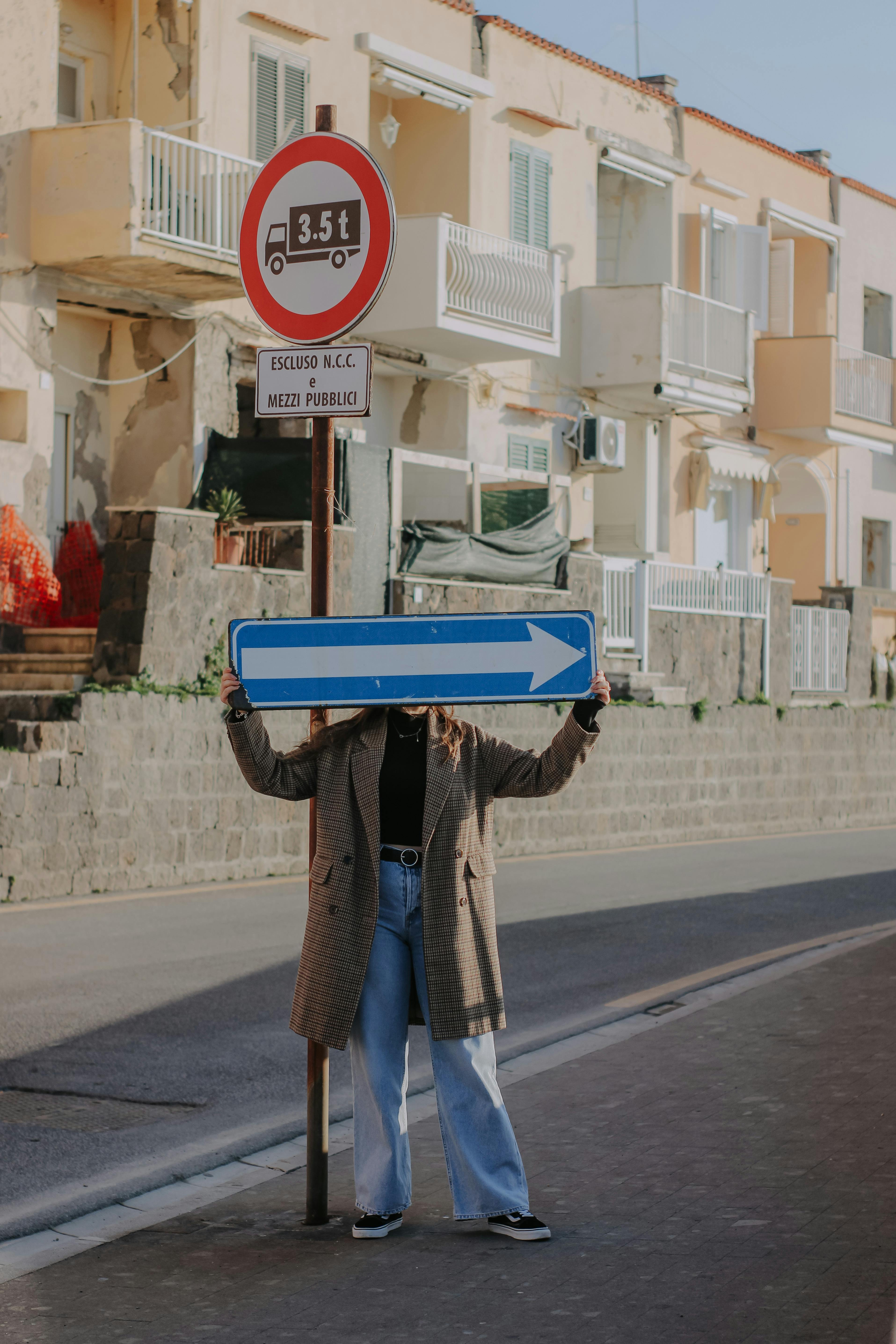A woman on a city street conceals her face with a directional arrow sign.