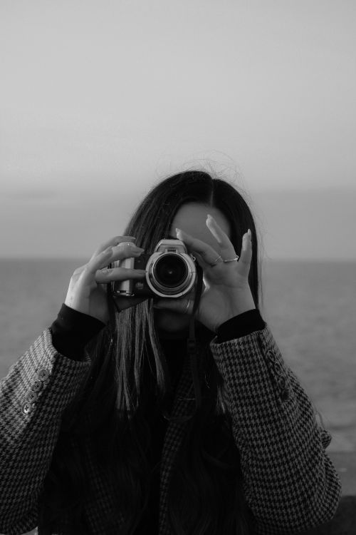 Free A woman capturing the moment with a camera by the sea, in a black and white portrait. Stock Photo