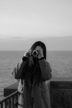 Black and white photo of a woman taking a picture with a vintage camera by the sea.
