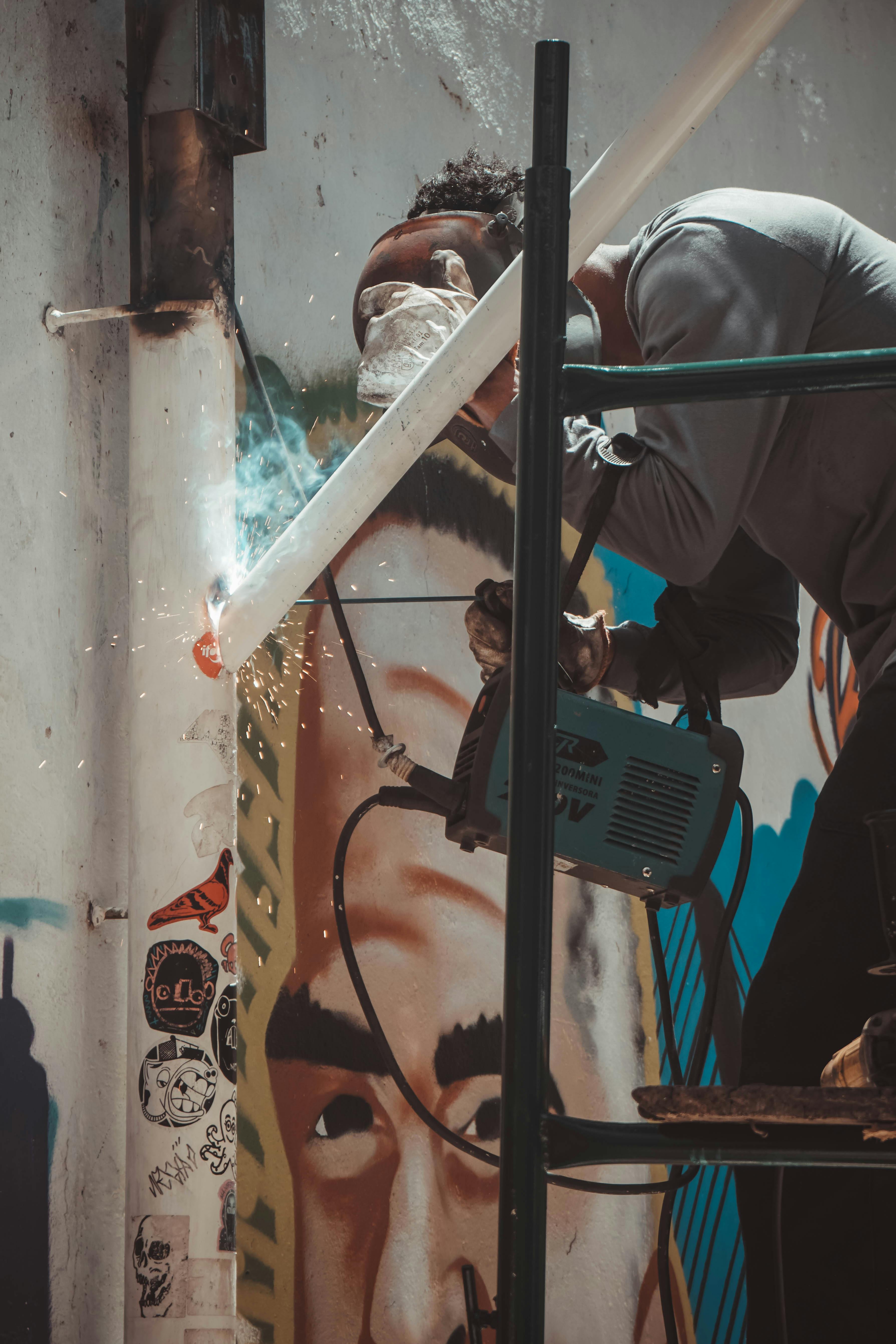 An artist welding a metal pipe to a wall in front of a vibrant mural, creating sparks.