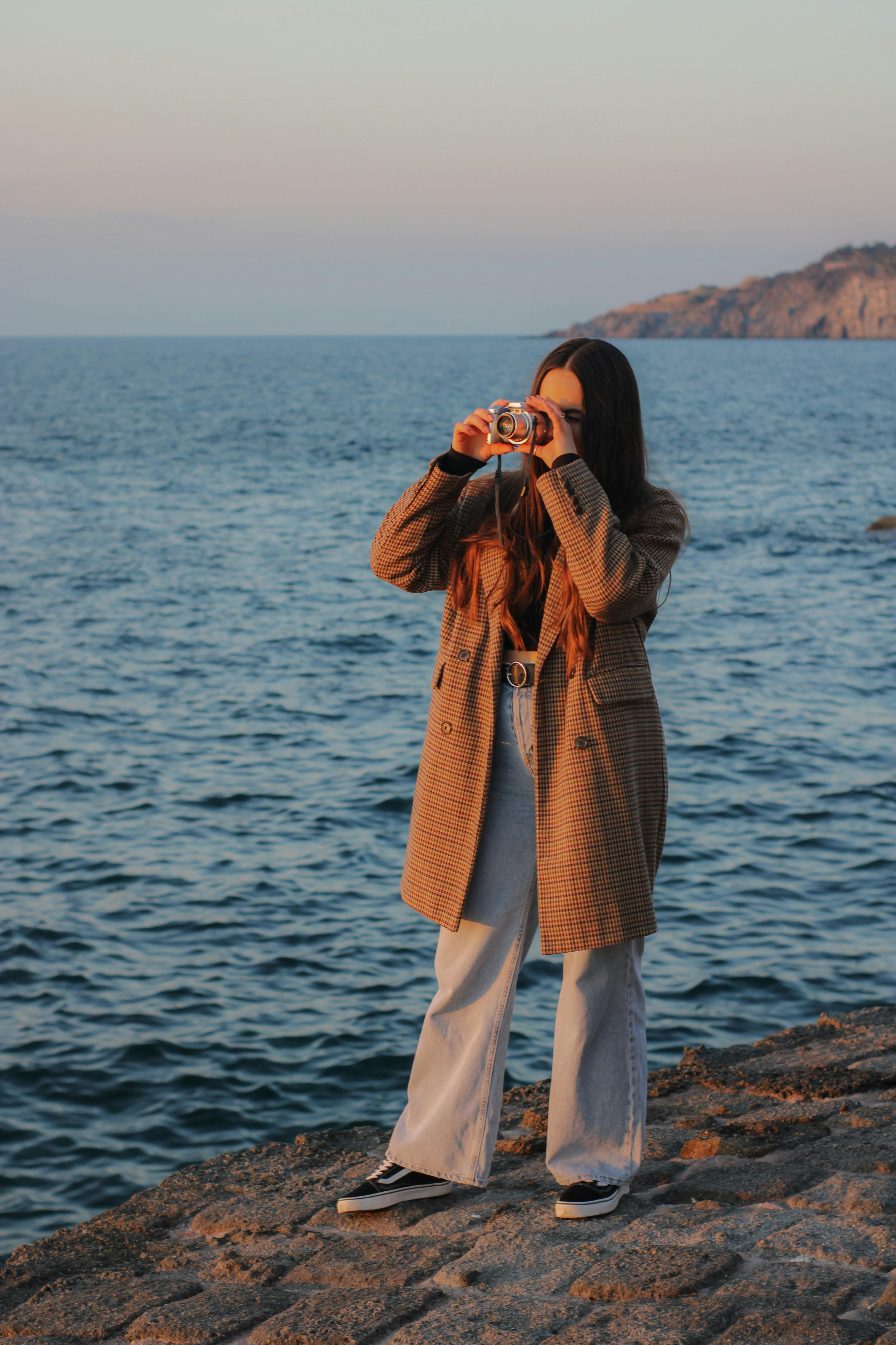 A young woman in a trench coat taking photos by the sea during sunset.