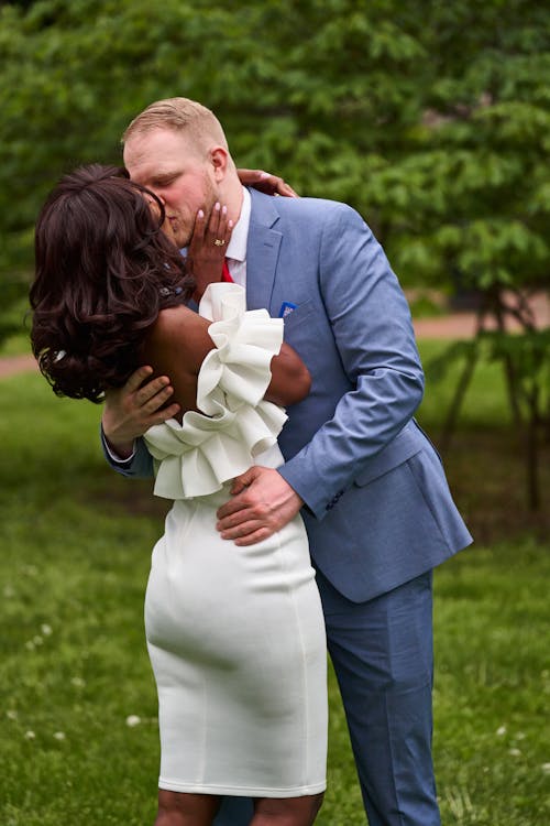 Man Wearing a Suit Kissing a Woman Wearing a White Dress with Ruffles in a Park