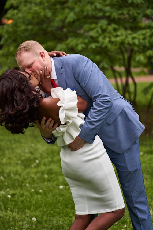 Man Wearing a Suit Kissing a Woman Wearing a White Dress with Ruffles in a Park