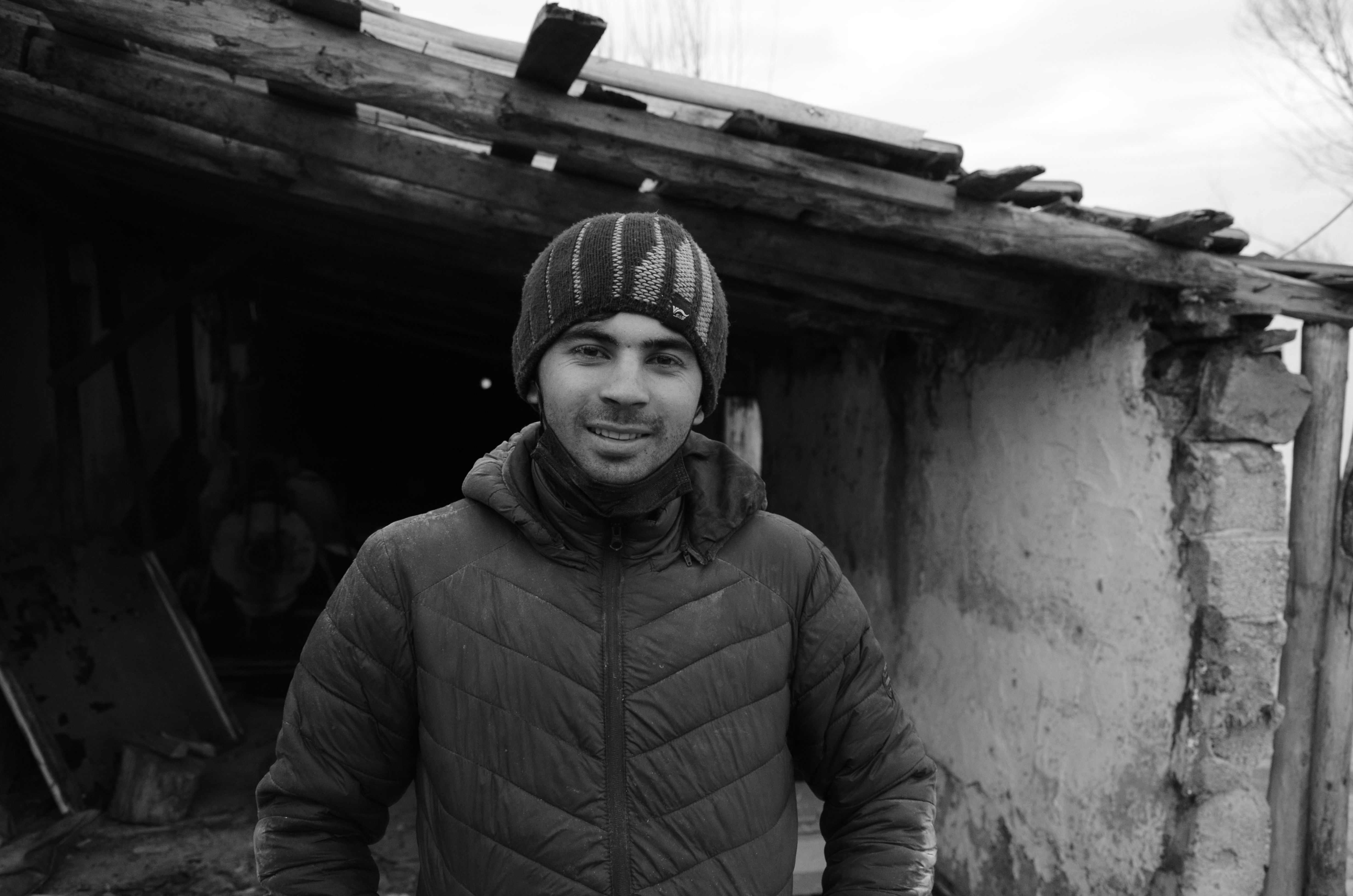 Black and white portrait of a man smiling in front of a dilapidated structure.