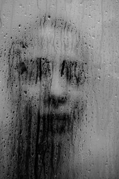 Mysterious black and white portrait of a woman seen through wet glass with water droplets.