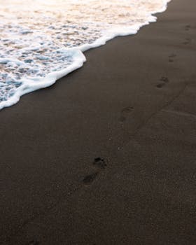 Tranquil scene of footprints on a black sand beach with gentle ocean waves.