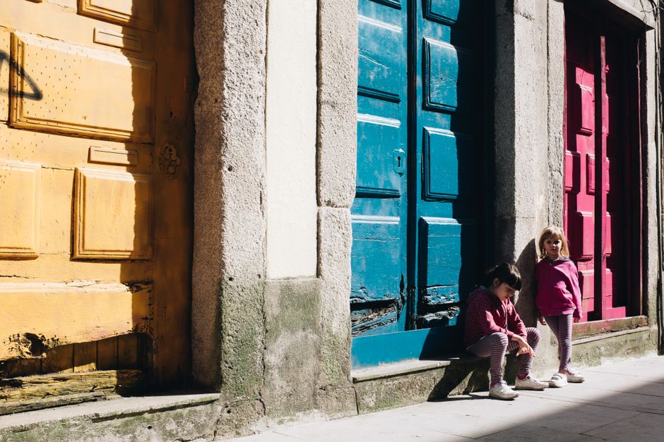 Two children sit outside vibrant colored doors in a sunny urban environment, capturing warmth and playfulness.