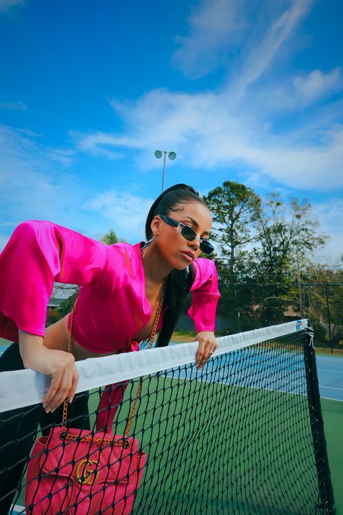 Free Stylish woman in sunglasses and pink blouse leaning on a tennis net outdoors. Stock Photo