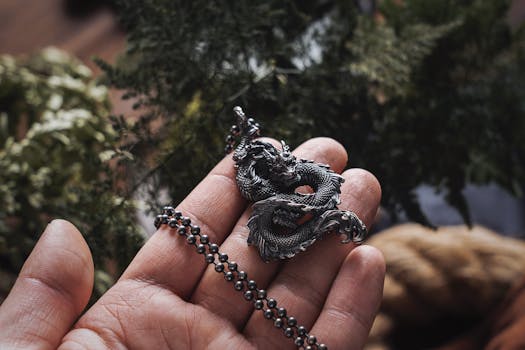 Close-up of a hand holding an ornate Chinese dragon necklace, surrounded by greenery.