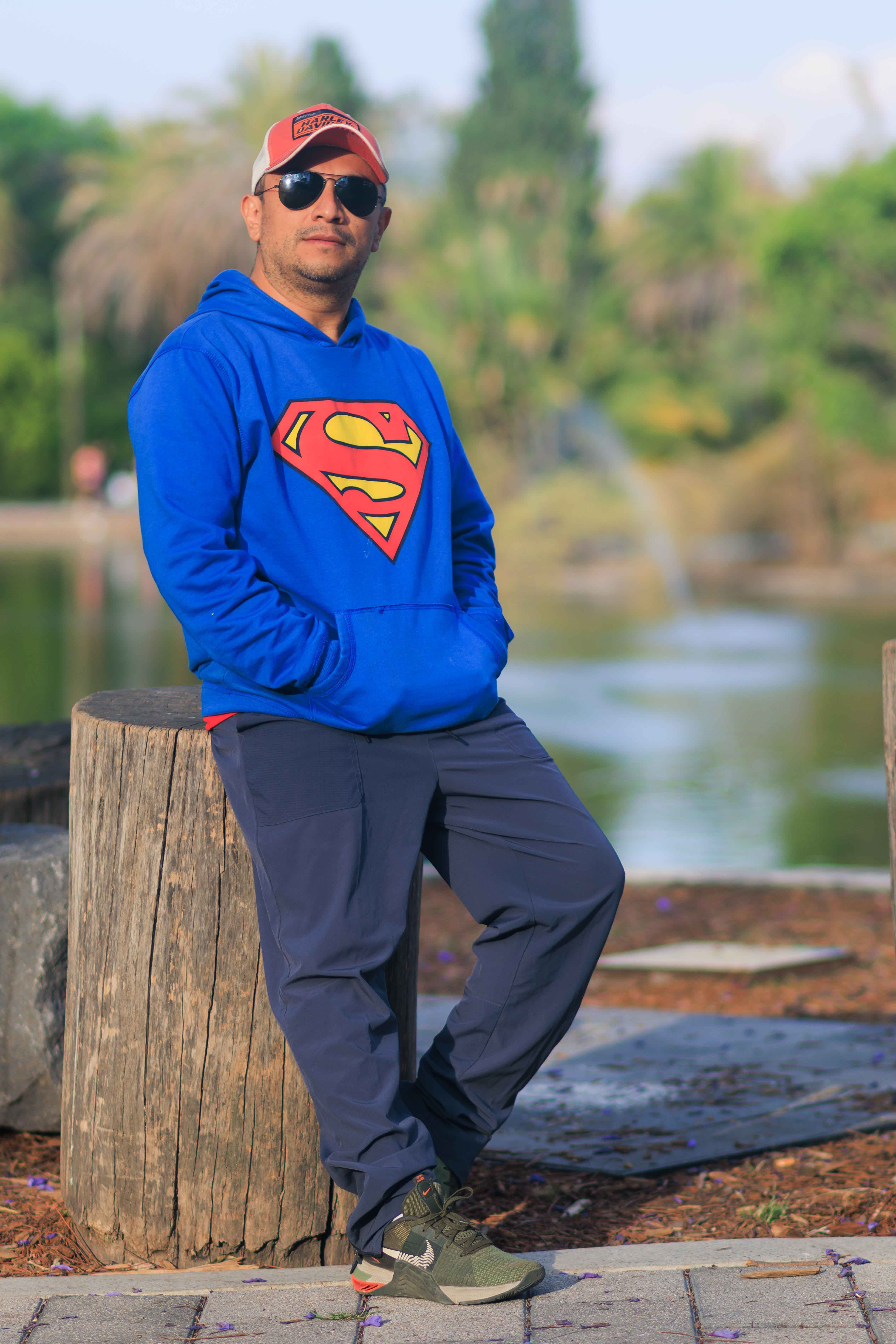 Man in blue Superman hoodie and cap stands casually outdoors near a lake.