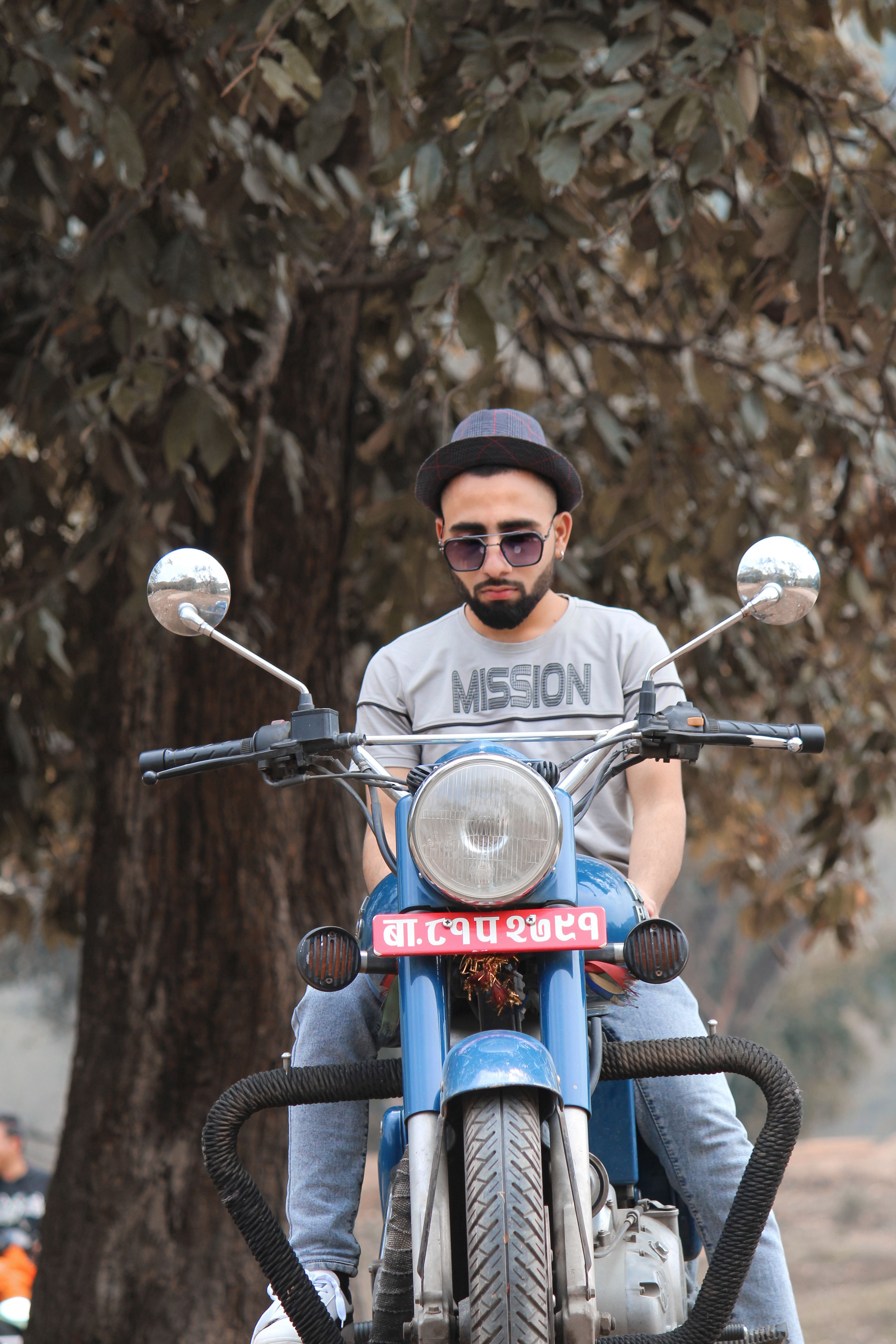 A trendy man wearing sunglasses rides a motorcycle in a natural setting.